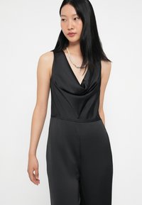 Macacão de satin preto com um decote em V profundo, frente drapeada e corte ajustado. Apresenta design sem mangas e silhueta de perna estreita.