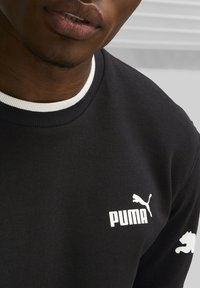 Czarna bluza z prążkowanym białym kołnierzem, białym logo Puma oraz charakterystycznym sylwetką kota na prawym rękawie. Gładka faktura tkaniny.