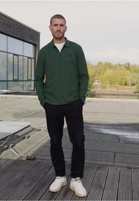 Zelená polo košile s dlouhým rukávem a čtvrtzipem, černé kalhoty a bílé tenisky s zelenými akcenty, stojící na střešní terase.