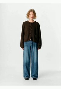 Brun strikket cardigan med frontknapper og lommer, kombinert med vidde blå denimjeans. Teksturen virker myk og koselig.