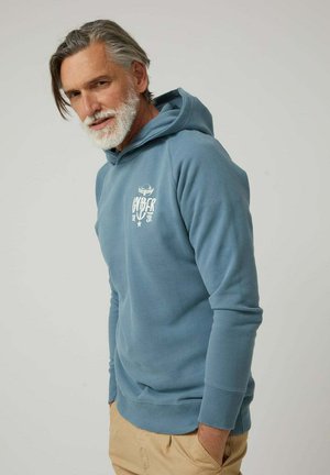 Hombre de mediana edad con barba gris, vestido con sudadera con capucha azul y pantalones beige, de pie con las manos en los bolsillos frente a un fondo liso.