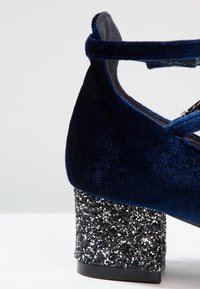Gros plan d'une chaussure en velours bleu marine avec un talon bloc recouvert de paillettes noires et argentées, avec une bride de cheville assortie.