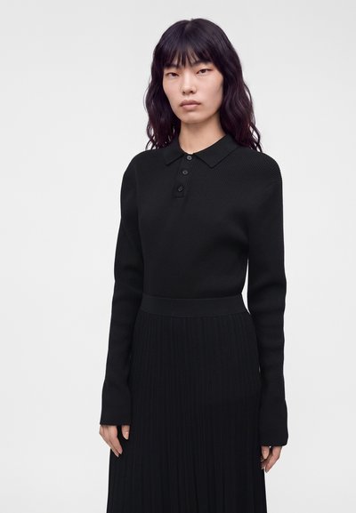 Femme aux longs cheveux foncés portant une chemise noire à manches longues avec col et une jupe plissée, debout devant un fond clair uni.