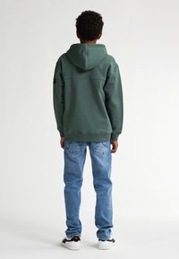 Groene hoodie met een relaxte pasvorm, gecombineerd met lichtblauwe jeans. De hoodie heeft een naaddetail en ribgebreide manchetten.