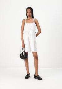White tiered dress with thin straps, textured fabric, and a relaxed fit. Paired with a black circular handbag and black loafers.