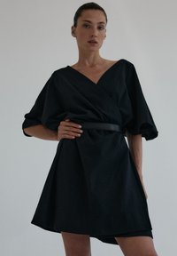 Czarna sukienka w stylu wrap z krótkimi, bufiaste rękawami i głębokim dekoltem w kształcie V. Posiada zawiązywany pas i rozkloszowany dół, wykonana z teksturowanej tkaniny.