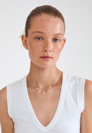 Mladi žena plavih očiju i zalizane kose, nosi bijelu majicu sa V-izrezom i srebrnu ogrlicu s privjeskom morske zvijezde na jednobojnoj pozadini.