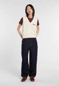 Chaleco de punto color crema con escote en V, dobladillo acanalado y detalles en burdeos, sobre una camiseta de cuello redondo burdeos; combinado con unos pantalones de mezclilla anchos de color azul oscuro.