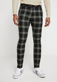 Muž v černobílých kostičkovaných slim-fit kalhotách, béžovém trikotu s dlouhým rukávem a bílých teniskách s tmavým hvězdicovým logem.