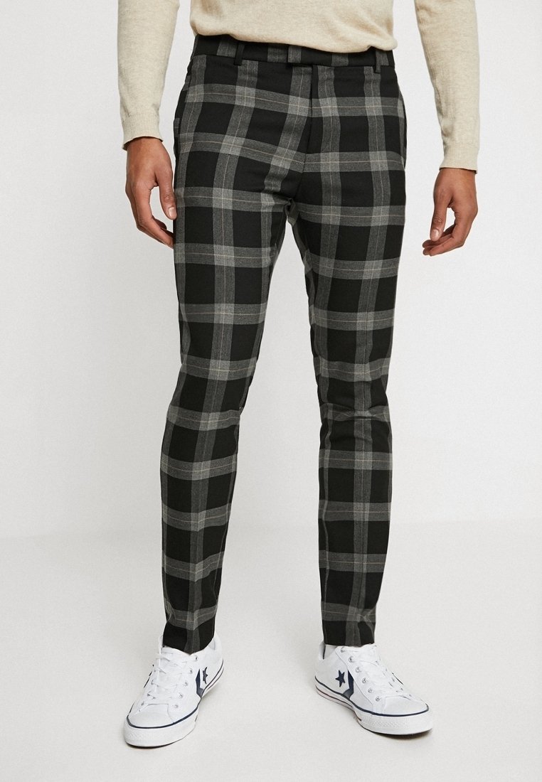 Muž v černobílých kostičkovaných slim-fit kalhotách, béžovém trikotu s dlouhým rukávem a bílých teniskách s tmavým hvězdicovým logem.