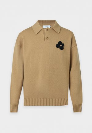 Jeune homme portant un pull en tricot beige avec un détail fleur noire, un pantalon ample foncé et des mocassins bicolores, debout les mains dans les poches.