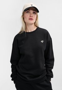 Svart sweatshirt med rund halsringning och ribbade ärmslut. Har en liten vit grafisk design på bröstet. Kombinerad med en beige basebollkeps.