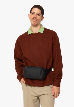 Sac banane noir avec fermeture éclair, porté par-dessus une chemise à manches longues marron avec une chemise à col verte en dessous, associé à un pantalon beige clair.