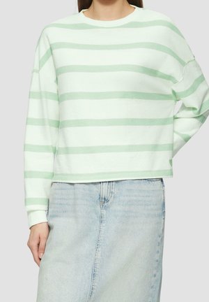 Person iført en lysegrøn og hvid langærmet top med vandrette striber og en lyseblå denims nederdel, står med den ene hånd på hoften.