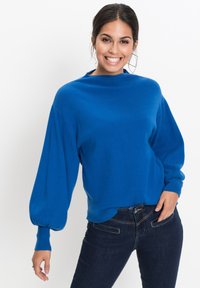 Maglione blu lavorato a maglia con collo alto e maniche a palloncino. Indossato con jeans scuri, caratterizzati da una texture liscia e una vestibilità rilassata.