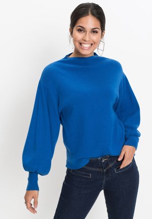 Pull en tricot bleu avec un col haut et des manches ballons. Porté avec un jean foncé, offrant une texture lisse et une coupe décontractée.