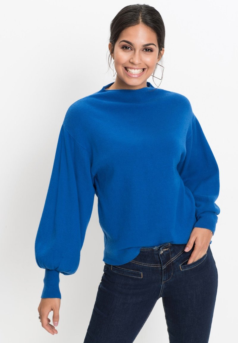 Maglione blu lavorato a maglia con collo alto e maniche a palloncino. Indossato con jeans scuri, caratterizzati da una texture liscia e una vestibilità rilassata.