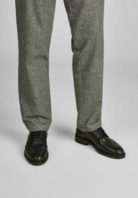 Schwarze Leder-Brogue-Schuhe mit durchbrochener Detailgestaltung, kombiniert mit hellgrauen strukturierten Hosen, die einen maßgeschneiderten Schnitt zeigen.