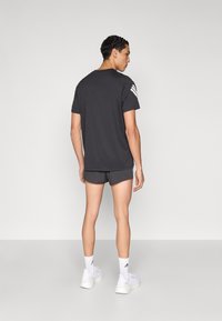 T-shirt de sport noir à manches courtes avec des accents de bandes blanches, porté avec un short foncé et des baskets blanches, sur un fond neutre.