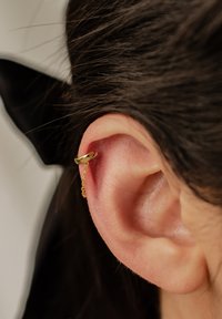 Goldene Creolen mit Kettendetail, auf einem Nahaufnahme eines Ohres präsentiert. Haare verdecken teilweise das Ohrläppchen für einen Kontrast.