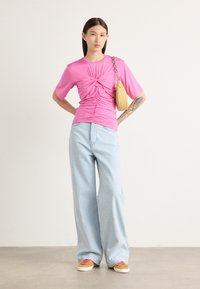 Haut rose ajusté avec des détails froncés, associé à un jean large bleu clair et des baskets colorées. Un petit sac jaune pend à l'épaule.