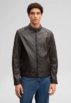 Mann steht, trägt eine schwarze Lederjacke mit Reißverschluss und gesteppten Schultern sowie blaue Jeans, blickt nach vorne vor einem hellen, schlichten Hintergrund.