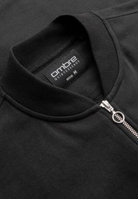 Chaqueta negra con cremallera y cuello y puños acanalados, que presenta una cremallera plateada y una etiqueta que dice "ombre QUINTESSENCE" en talla M.