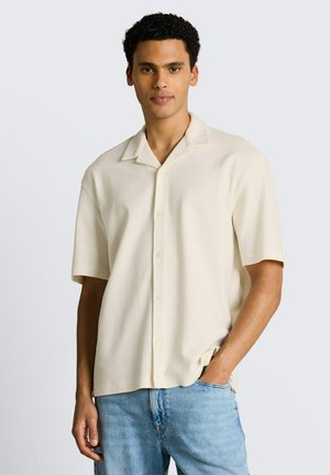 Junger Mann trägt ein cremefarbenes kurzärmeliges Hemd mit Knöpfen und hellblaue Jeans, steht mit einer Hand in der Tasche vor neutralem Hintergrund.