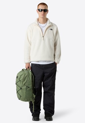 Fleece pullover in off-white, zip neck, black logo, paired with dark pants and a green backpack featuring a mesh pocket and bungee cords.