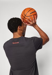 Juoda sportinė marškinėliai su mažu raudonu "Reebok" logotipu ant nugaros, laikomi virš galvos oranžiniu krepšiniu, vientisas fonas.