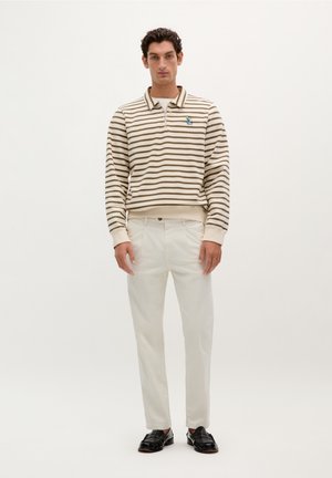 Gestreepte, beige en bruine pullover met een kraag en halve rits, gecombineerd met witte broek en zwarte instappers. Opvallend geborduurd detail.
