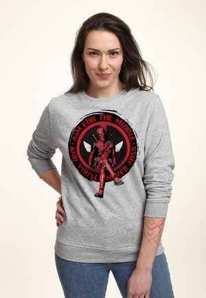 Kvinde med langt mørkt hår iført en grå sweatshirt med Deadpool-grafik og rød tekst, stående med den ene hånd på hoften.
