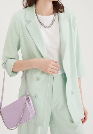 Femme portant un blazer vert menthe et un pantalon avec une chemise blanche, un collier chaîne argenté, et portant une petite pochette lavande texturée avec une bandoulière chaîne.