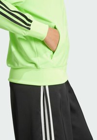Neon-grüner Sportpullover mit schwarzen Streifen an den Ärmeln, gerippten Bündchen und einer Reißverschlusstasche. Kombiniert mit schwarzen Shorts, die weiße Streifen haben.