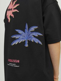 Juodas medvilnės marškinėliai su rožiniu saule ir mėlynos palmių lapo grafika, dekoruoti auksiniais akcentais. Tekste yra "ORIGINALS" ir "NINETEEN NINETY" rožinės ir baltos spalvos.
