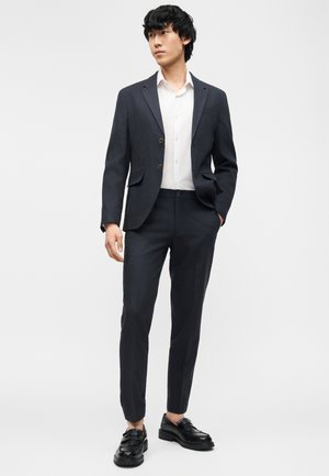 Mann trägt einen dunkel maßgeschneiderten Anzug mit weißem Hemd und schwarzen Loafers, steht mit einer Hand in der Tasche vor einem einfarbigen Hintergrund.
