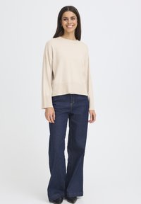 Bež pulover kratke dolžine z dolgimi rokavi, kombiniran s temno modrimi jeans hlačami širokih hlačnic, značilna za preprost dizajn in lahkotno teksturo tkanine.