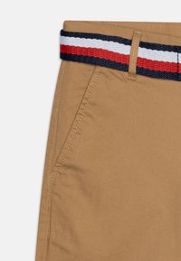 Tessuto khaki con finitura testurizzata, dotato di una cintura intrecciata blu, rossa e bianca. Include una tasca laterale e cuciture dettagliate.