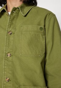 Olivgrön bomullsjacka med knappstängning, en patchficka och ett logomärke på bröstet. Texturerad tyg med en klassisk design.