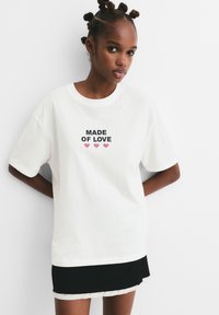 Mlada žena s frizurom u bantu čvorovima nosi bijelu majicu s natpisom "MADE OF LOVE" i tri ružičaste srca, u kombinaciji s crnom suknjom.
