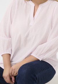 Jasnoróżowa bluzka o fakturze z kołnierzykiem mandarynkowym i elastycznymi rękawami w kształcie balonu, zestawiona z ciemnoniebieskimi dżinsami.