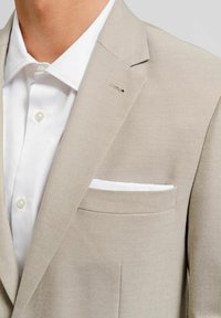 Ljusbeige kostymjacka med texturerad yta, har en vit bröstficka och en silvrig knappdetalj på slaget, tillsammans med en vit skjorta.