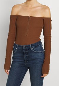 Hnědý žebrovaný off-shoulder bodysuit s dlouhými rukávy a předním zipem, kombinovaný s tmavě modrými džínami s vysokým pasem.