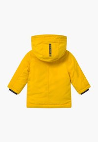 Veste isolante jaune avec capuche, dotée de bordures bleu marine aux poignets, de détails cousus et d'une étiquette au dos.