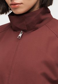 Κοντινό πλάνο ενός ατόμου που φοράει μπουφάν με ψηλό γιακά σε μπορντό χρώμα και ασημένιο φερμουάρ, με εμφανείς τα χείλη και το πηγούνι.