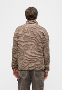 Άνδρας με καφέ και μπεζ φλις μπουφάν με σχέδιο και σκούρο παντελόνι cargo, εμφανιζόμενος από πίσω σε απλό ανοιχτό φόντο.