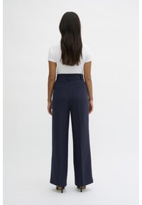 Navy wide-leg trousers with a high waist, two back pockets, smooth texture, and paired with a white short-sleeve top and heels.