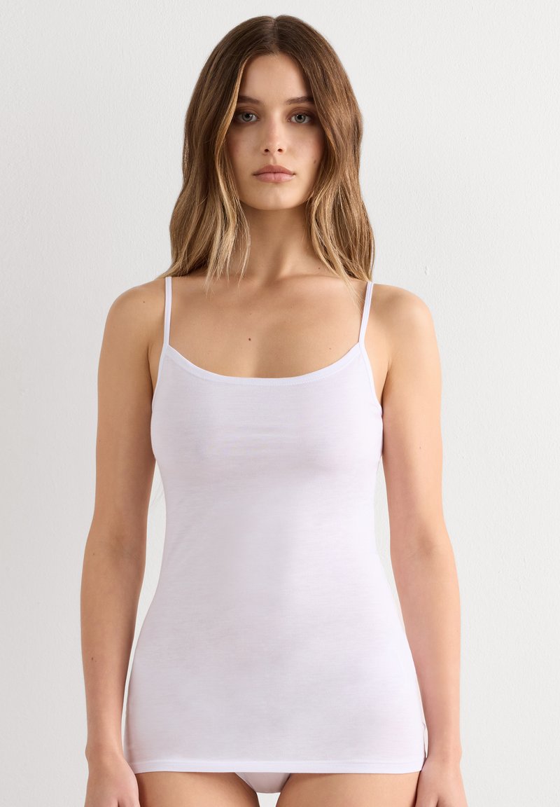 Young woman with long light brown hair wearing a fitted white spaghetti strap tank top against a plain background.