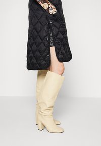 Kozačky z krémové umělé kůže do výšky kolen s robustním blokovým podpatkem. Hladký povrch a přiléhavý střih, s mírně volným okrajem nahoře.