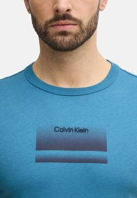 Camiseta de algodón turquesa con cuello redondo y un estampado del nombre de la marca "Calvin Klein" en negro sobre un fondo degradado.